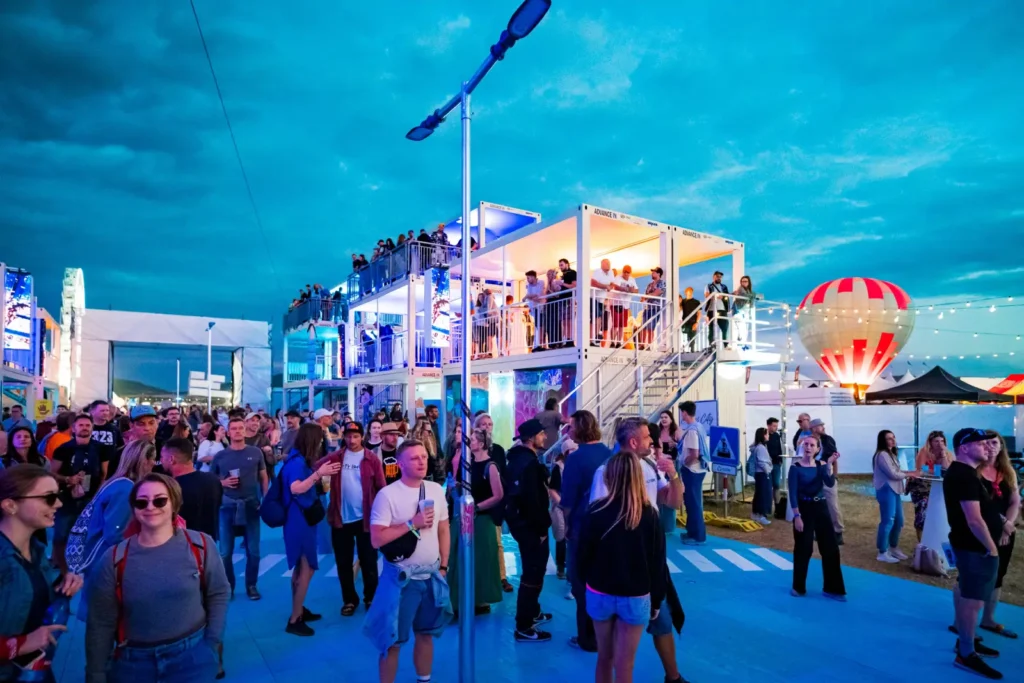 Festivalová ulica zóny IQOS Curious City na Pohode za súmraku – dav medzi kontajnerovými budovami, pouličná lampa a teplovzdušný balón v pozadí
