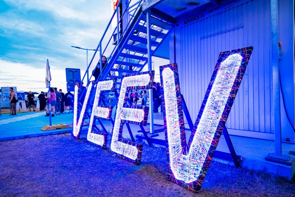 Veľký svietiaci nápis VEEV z farebných vlákien v zóne IQOS Curious City na Pohode za súmraku