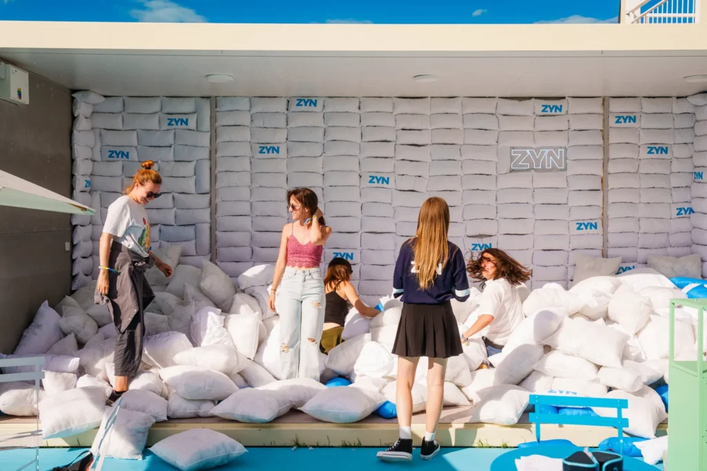 Aktivácia ZYN Pillow v zóne IQOS Curious City na Pohode – stena z brandovaných vankúšov a kopa vankúšov, v ktorých návštevníčky lovia ukrytý puk