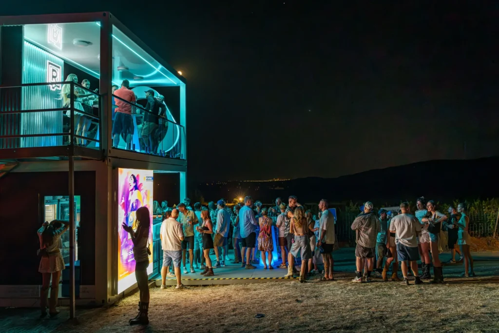 Nočný pohľad na nasvietenú kontajnerovú zónu IQOS Curious City na Ženy Víno Funk s davom návštevníkov